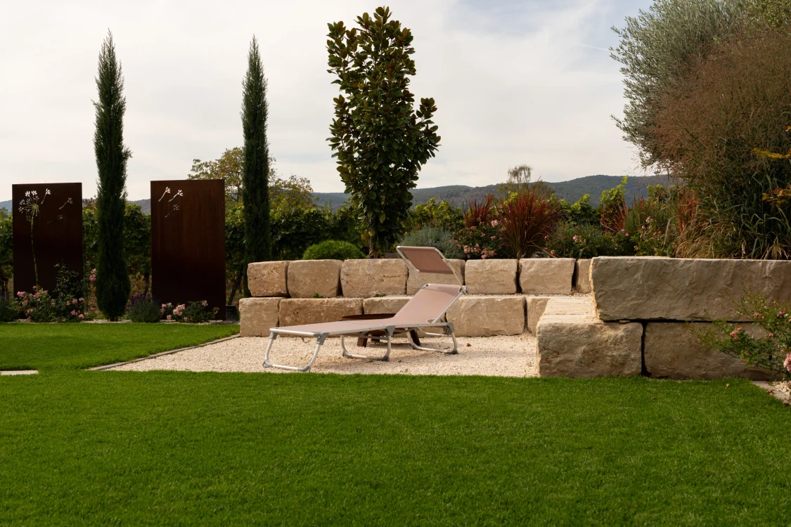 Lawn in front of the enclosed gravel area, with a garden lounger, slender woody plants and rust-finish metal features