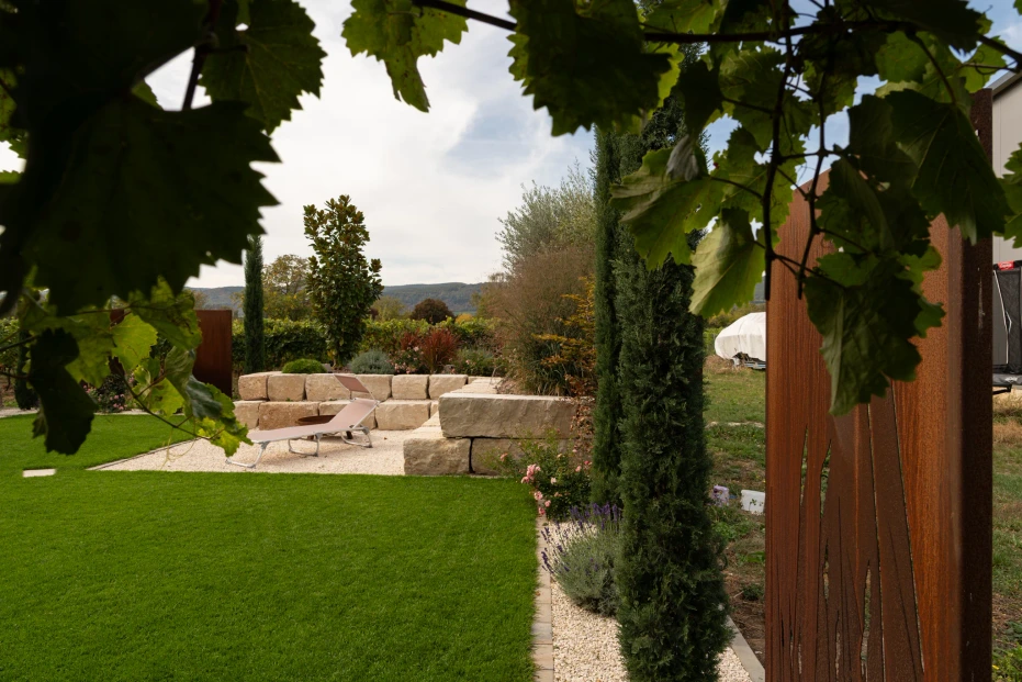 View through overhanging leaves towards the lawn and gravel area, with a rust-finish metal feature at the garden edge