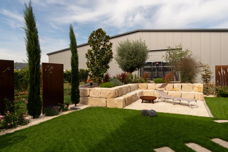 Gravel area with a lounger and fire bowl, surrounded by natural stone, woody plants and lawn